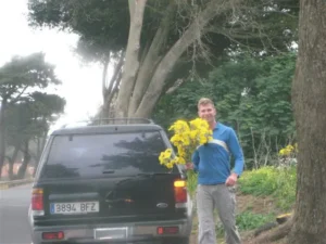 Mees jalutab pargis, hoides käes suurt kimp kollaseid lilli, taustal puud ja rohelus.